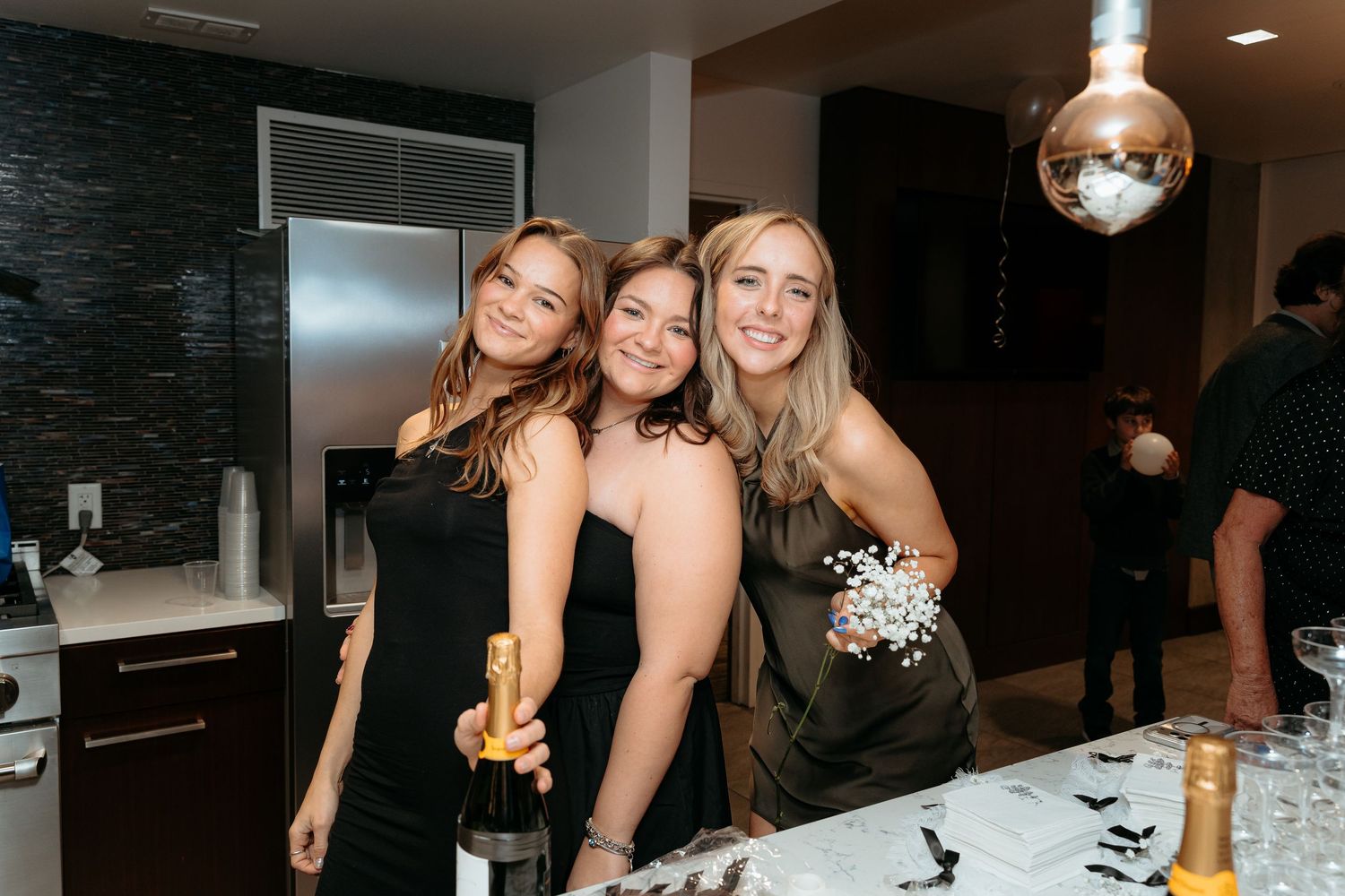 Three women in black dresses smile together at a New Year's Eve party with champagne and disco ball.