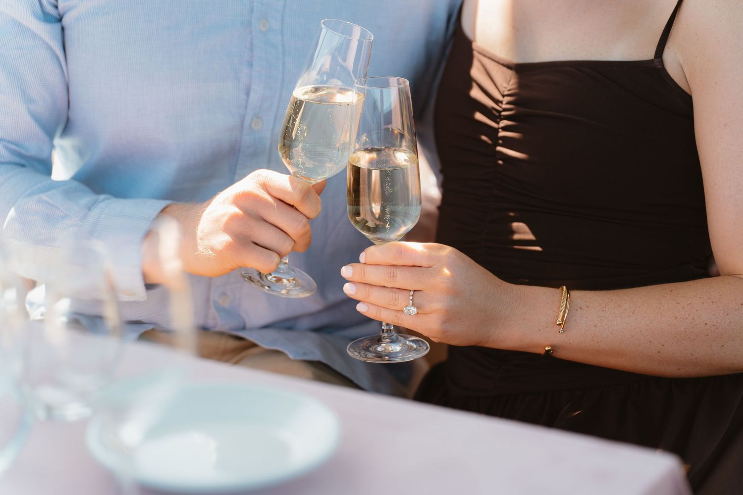 Couple clinking champagne glasses at romantic dinner, woman wearing engagement ring and gold bracelet.