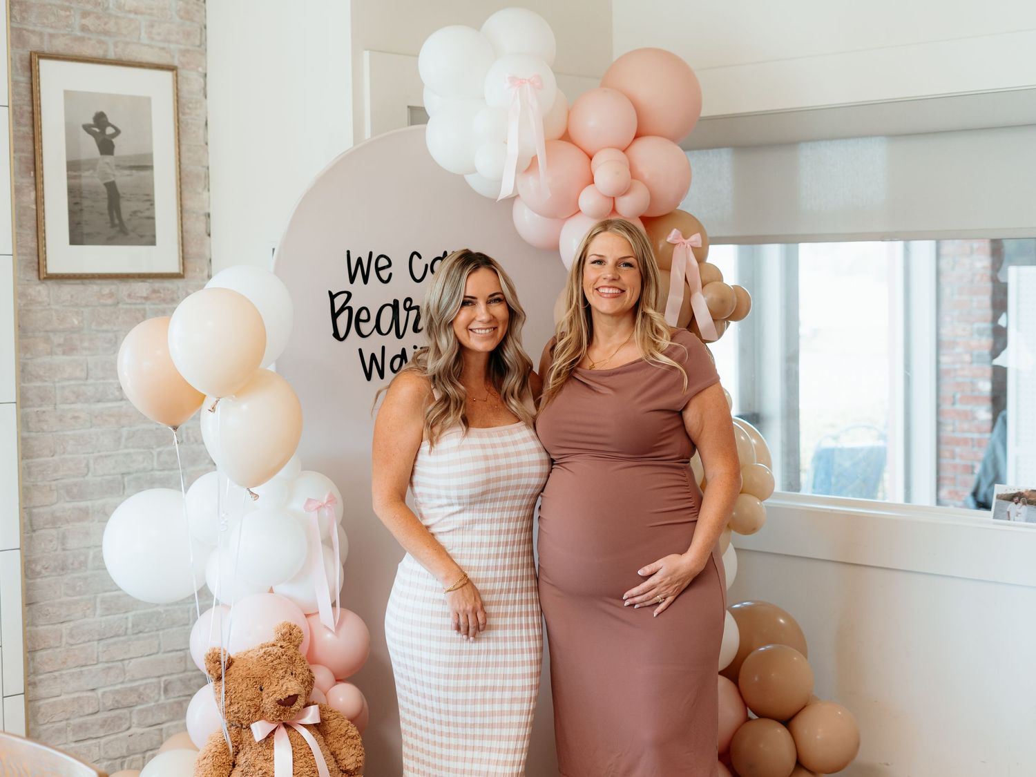 Two women pose at baby shower with pink and white balloon arch and teddy bear decorations.