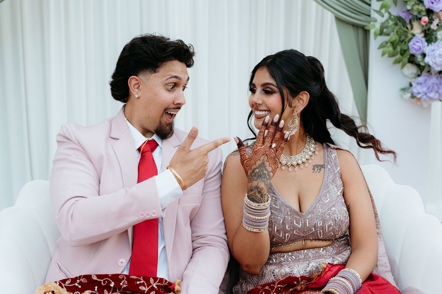 Couple in wedding attire, man in pink suit with red tie, woman in embroidered lehenga with henna hands, smiling together.