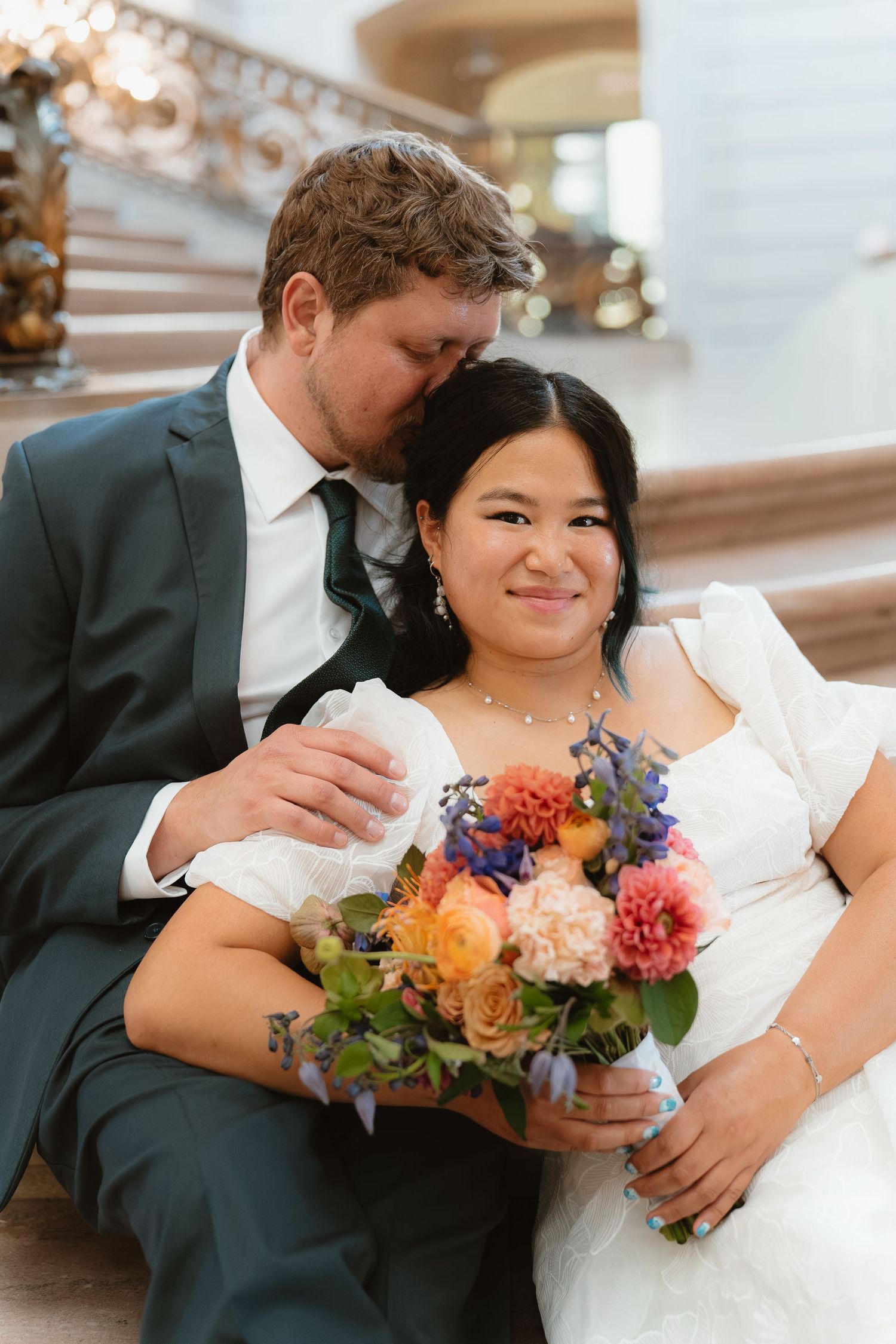 Bride and groom sit together on stairs, she holds a colorful bouquet of roses and dahlias in a white wedding dress.