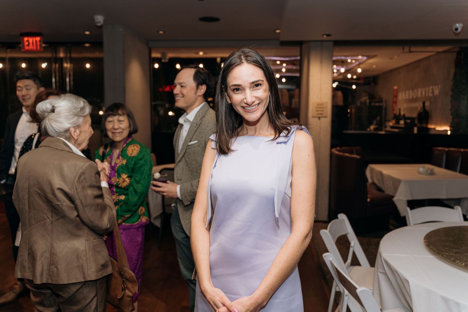 Major Milestone birthday where Woman in light blue dress smiles at indoor event with guests socializing in elegant restaurant background.