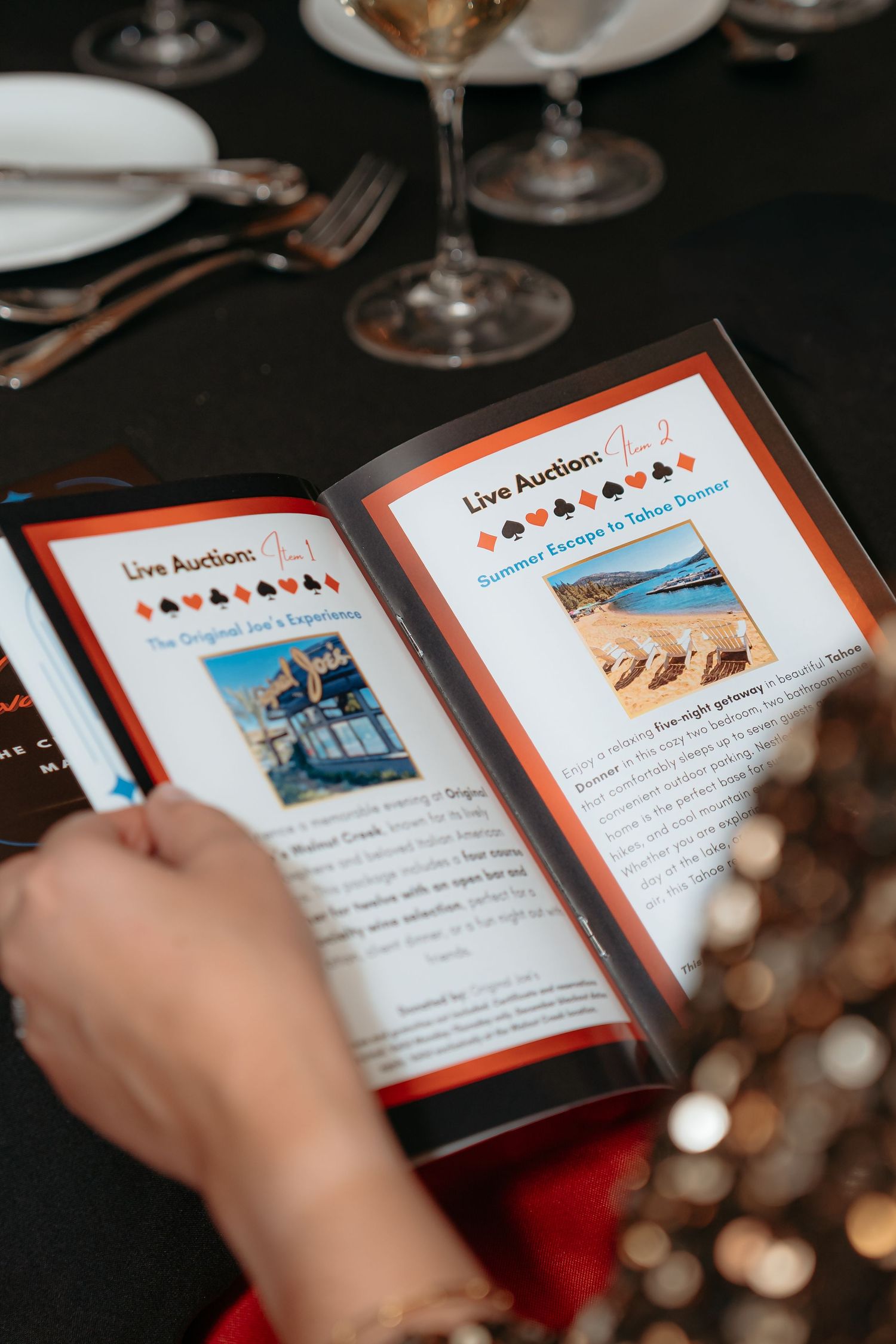 Hands holding a Live Auction booklet at a formal event table with glassware and black tablecloth.