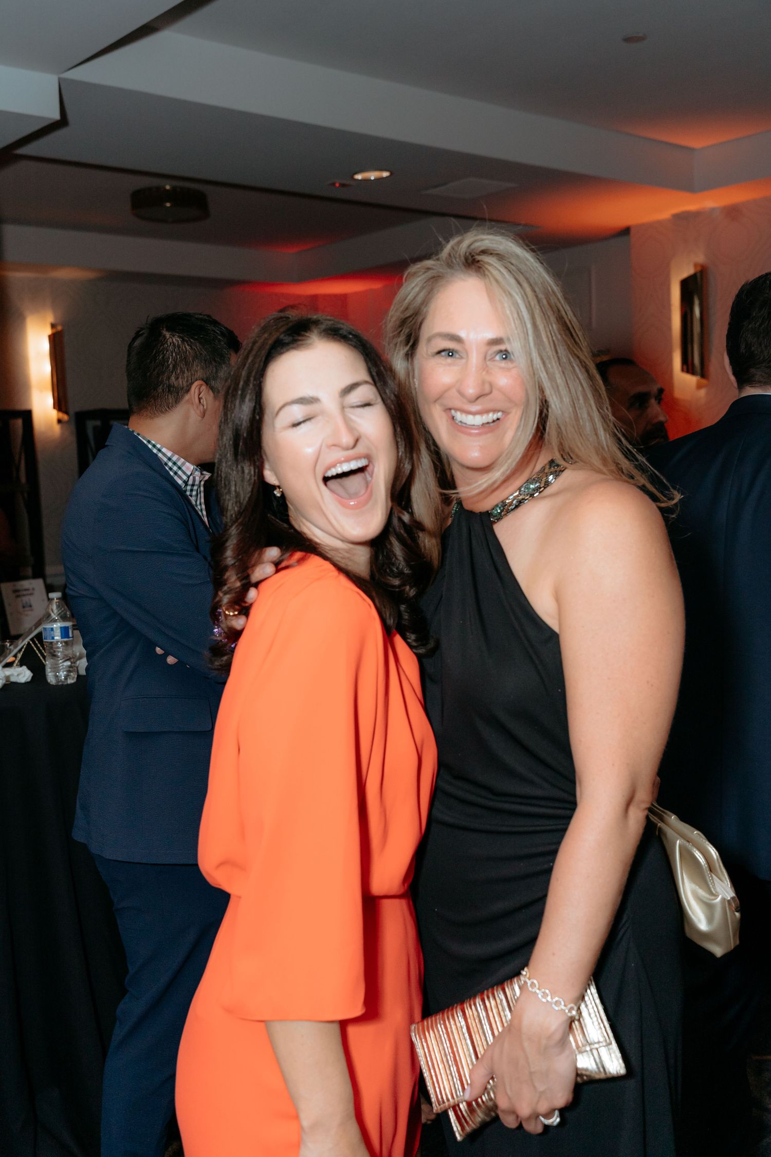 Two women laughing and posing together at a formal event, one in orange dress, one in black gown holding a clutch.