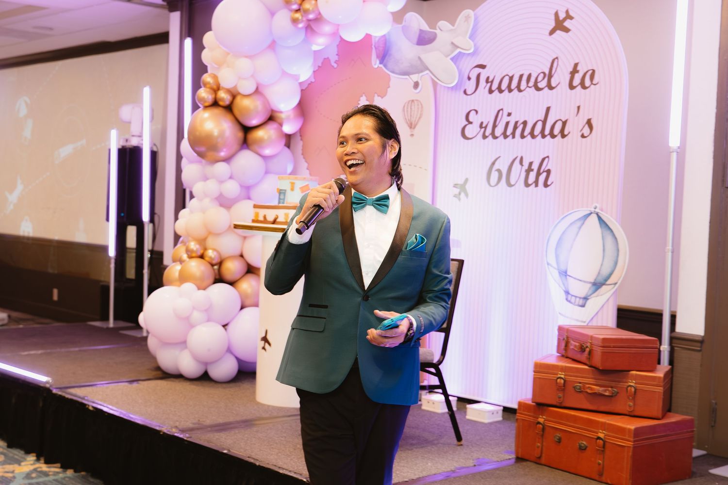 Man in teal blazer and bow tie speaks at microphone at Erlinda's 60th birthday travel-themed party.