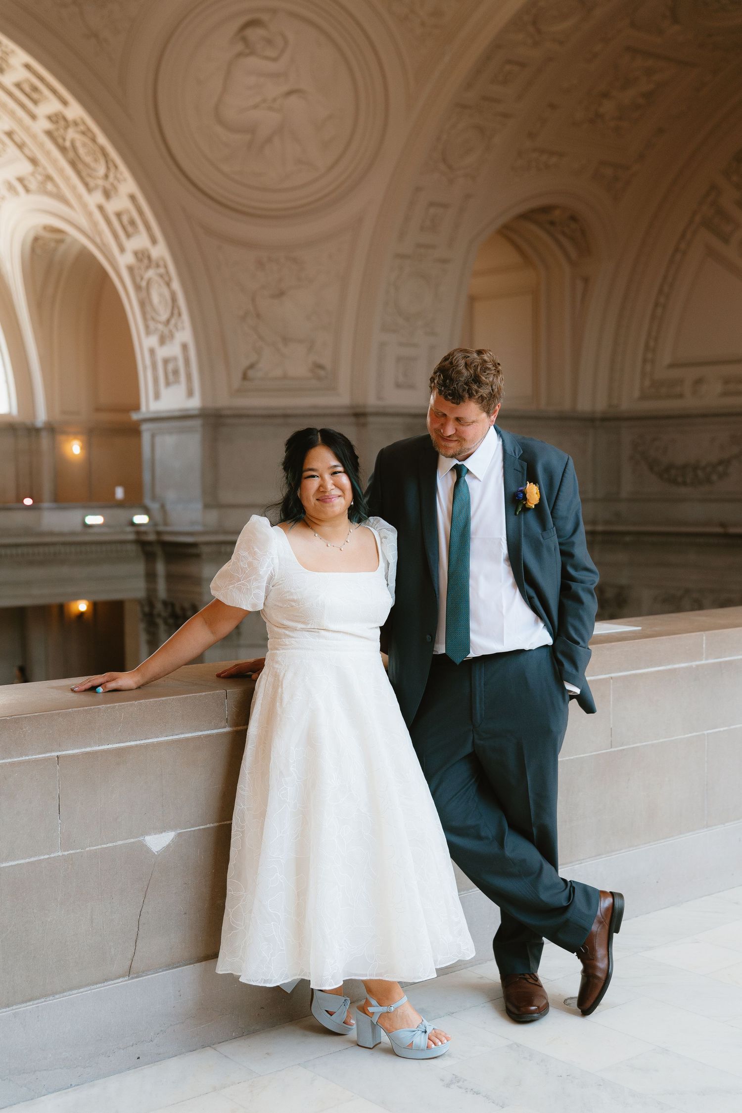 Bride in white midi dress and groom in dark suit pose in ornate San Francisco City Hall rotunda.