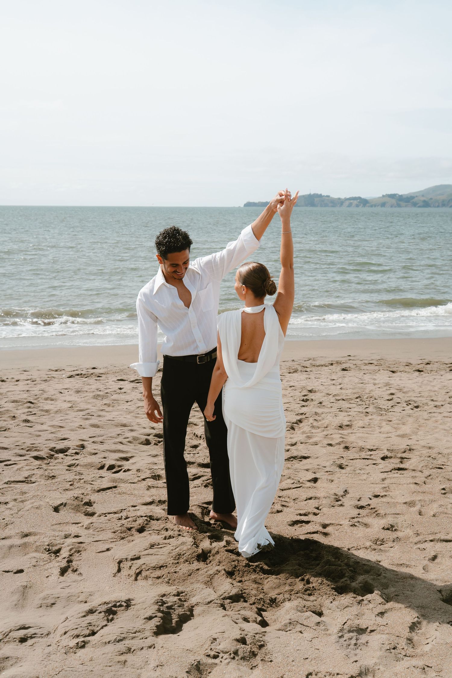 Couple dancing on a sandy beach, woman in white backless gown, man in white shirt and black pants, ocean backdrop.