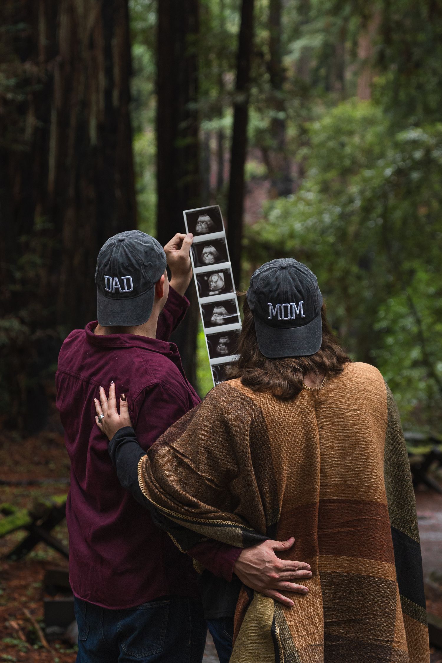 Couple wearing Dad and Mom hats holding ultrasound photos in a lush forest, announcing their pregnancy.
