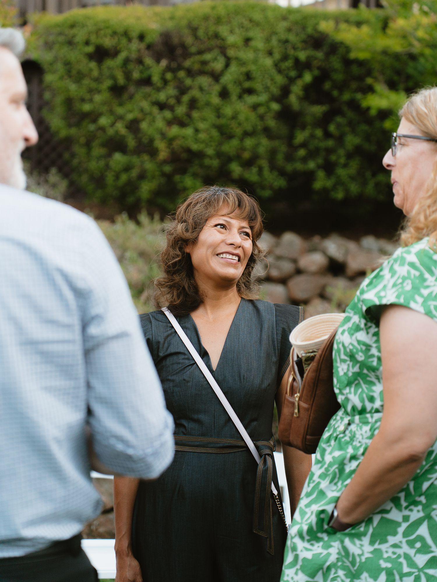 Woman in black dress smiling outdoors at a garden gathering with guests in casual attire.