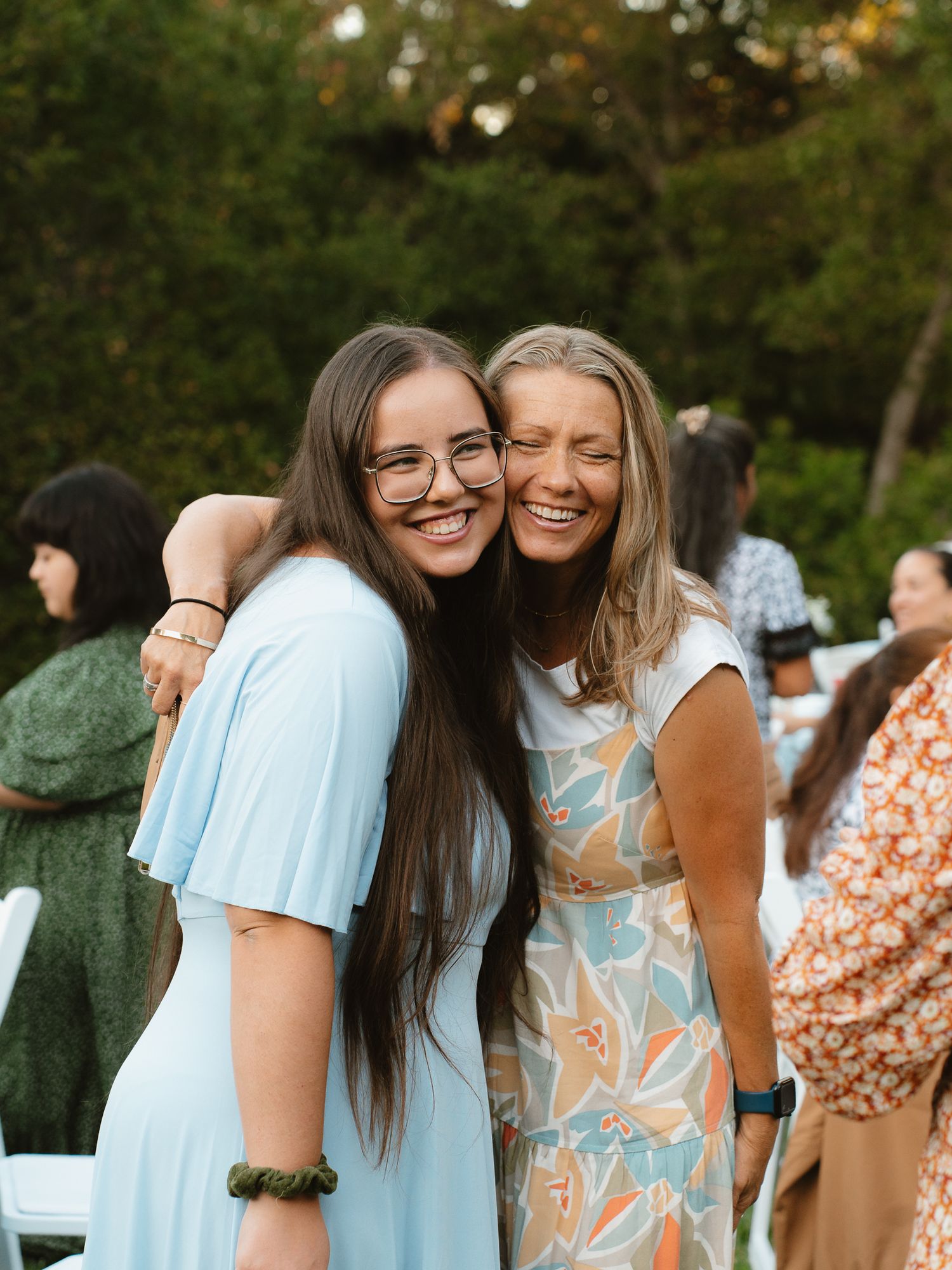 Two smiling women hugging outdoors at a garden event, one in a light blue dress and one in a floral dress.