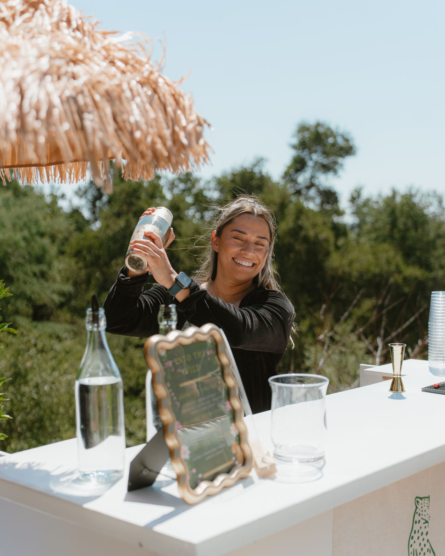 Bartending event Smiling female bartender pouring a cocktail at an outdoor bar under a thatched umbrella surrounded by lush greenery.