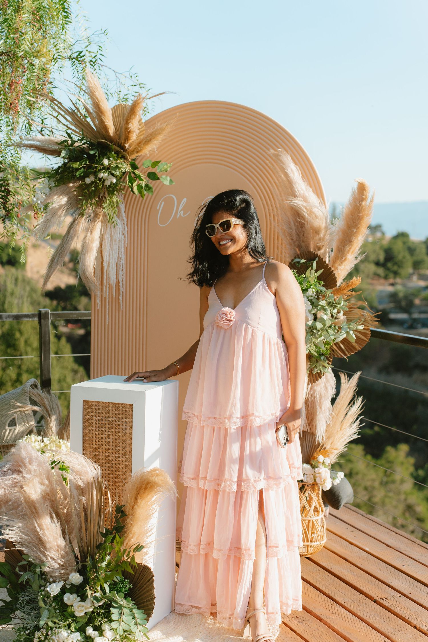 Gender reveal party in San Jose area where Woman in pink tiered dress posing by boho arch with pampas grass and greenery outdoors.