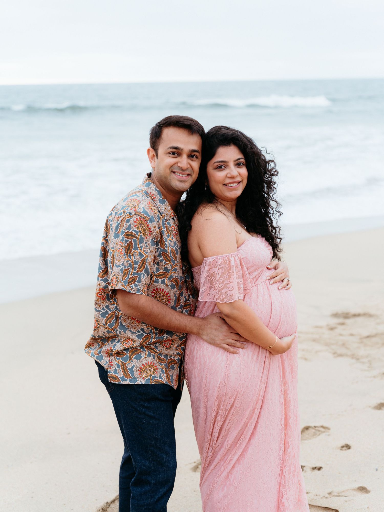 A pregnant couple poses for maternity session on a sandy beach, woman in pink maternity gown, man in floral shirt, ocean waves behind them.
