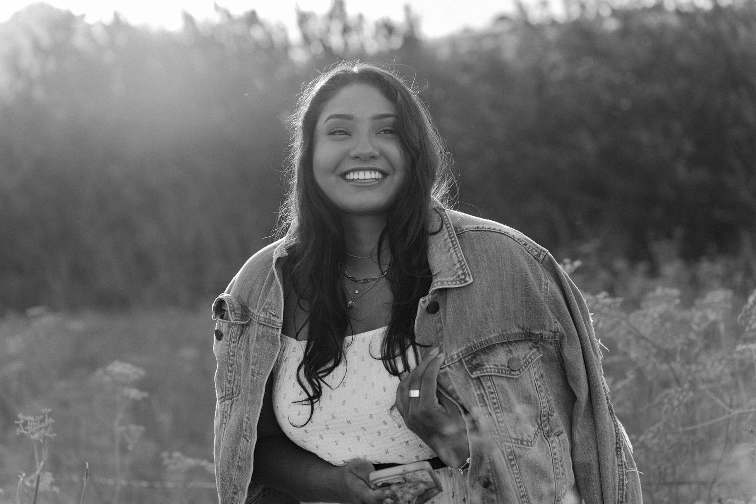 Black and white photo of a young woman smiling outdoors in a denim jacket, with sunlight and wildflowers behind her.