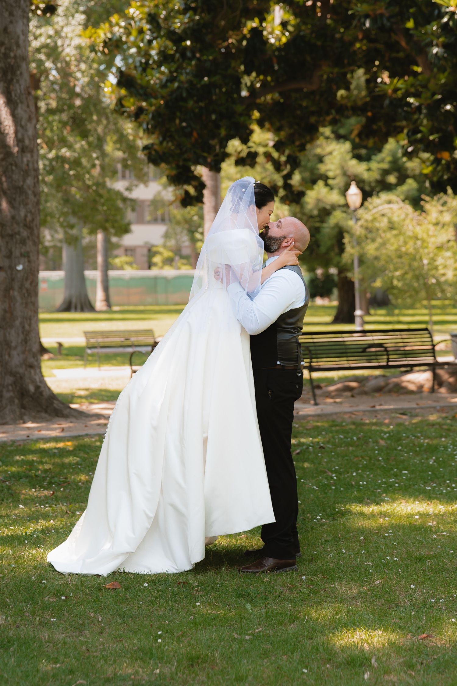 Groom lifting bride for a romantic kiss in a lush park, bride wearing white gown and veil.