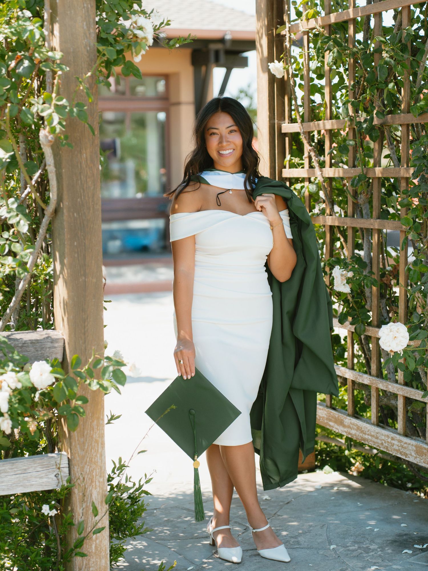 Graduate in white dress holding green cap and gown under floral garden arbor.
