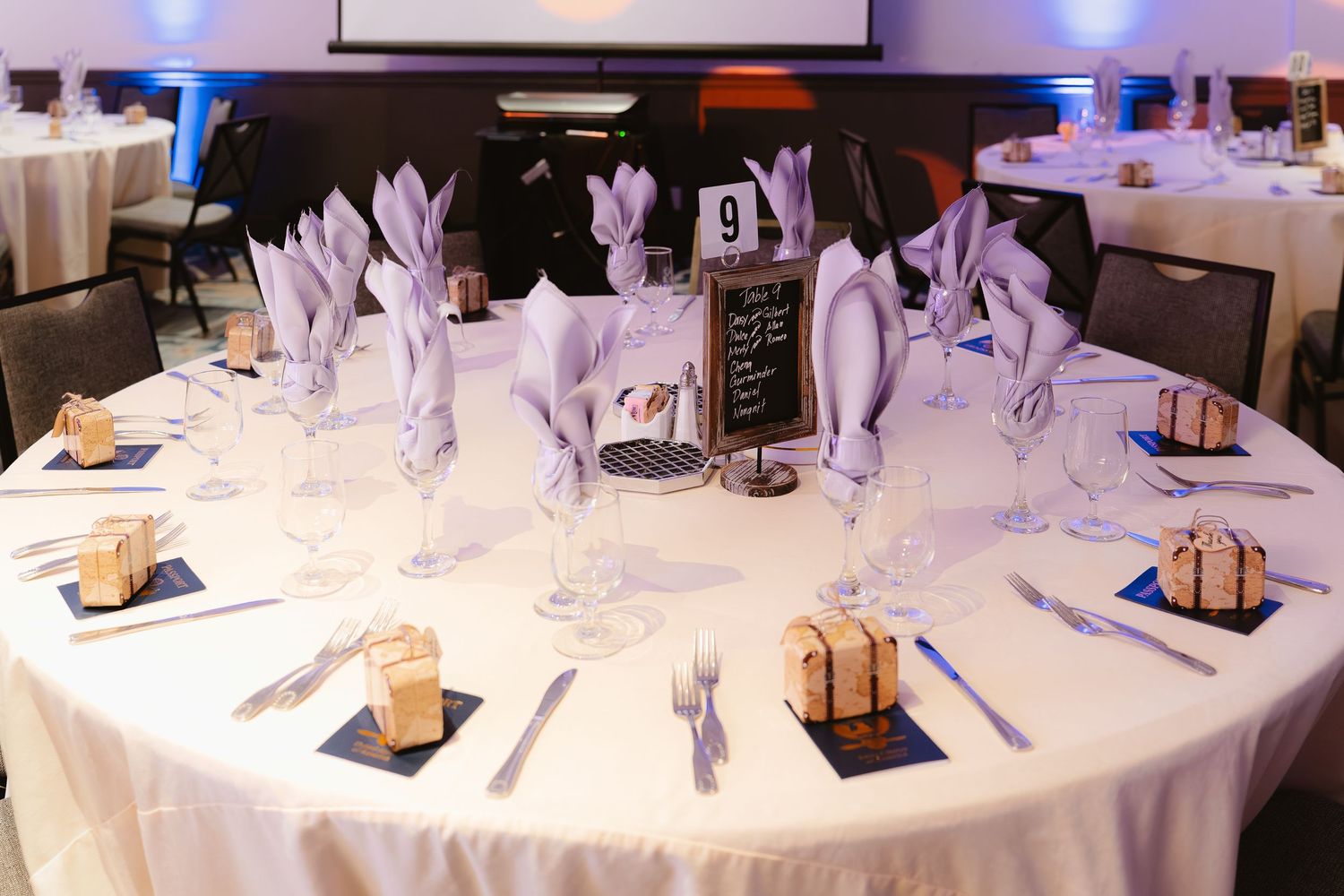 Elegant wedding reception table setting with lavender napkins, crystal glasses, and small gift boxes on a white tablecloth.