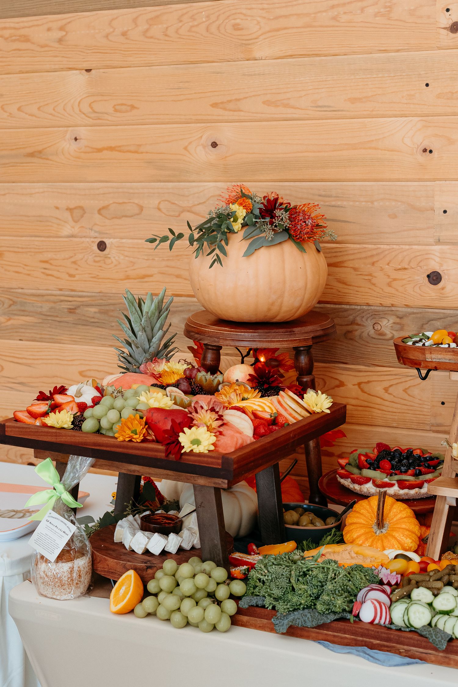 Elegant fall harvest charcuterie display with fruits, vegetables, pumpkins, and autumn flowers on a wooden tiered stand.