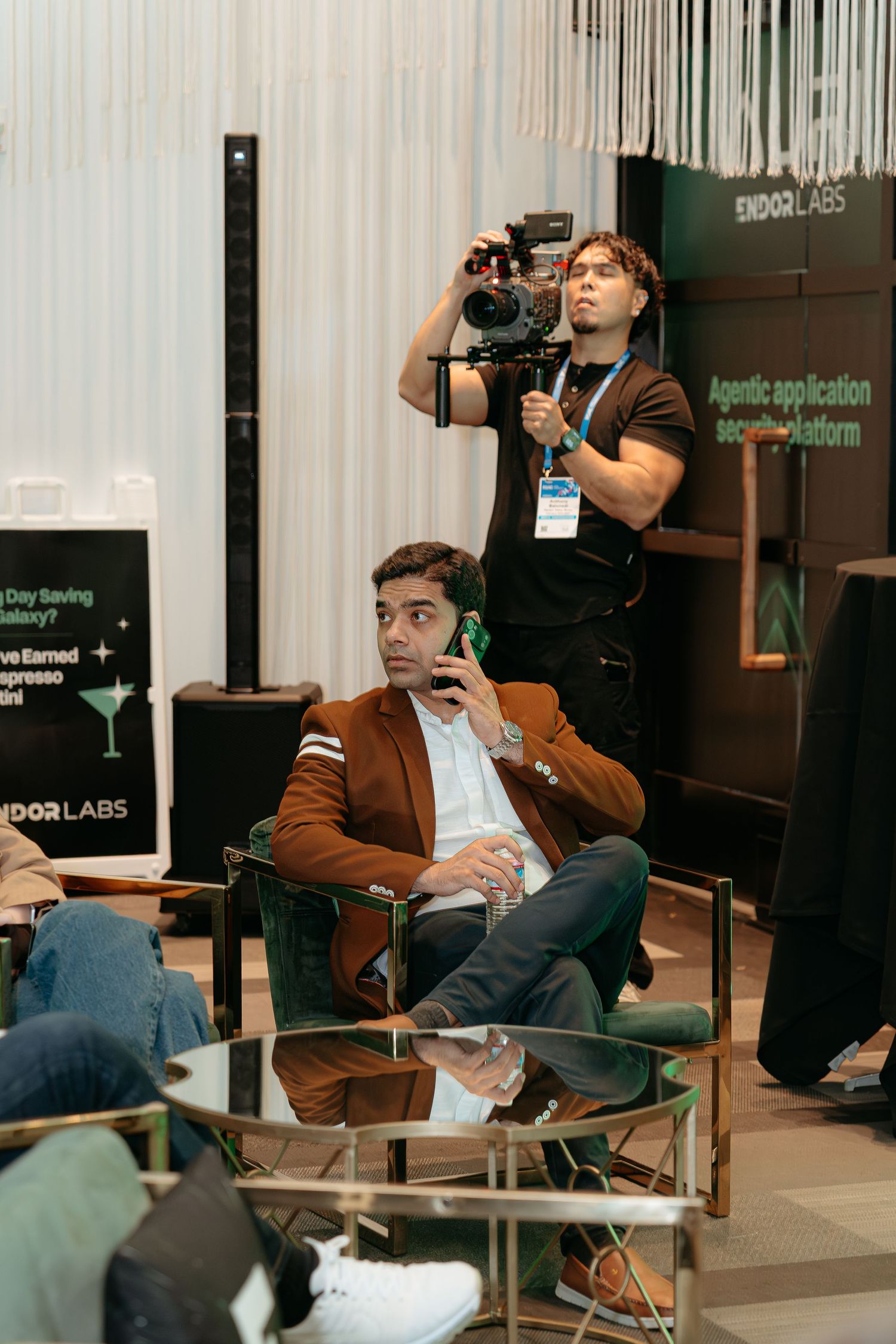 A man in a brown blazer sits on a chair while a videographer films at an Endor Labs event.
