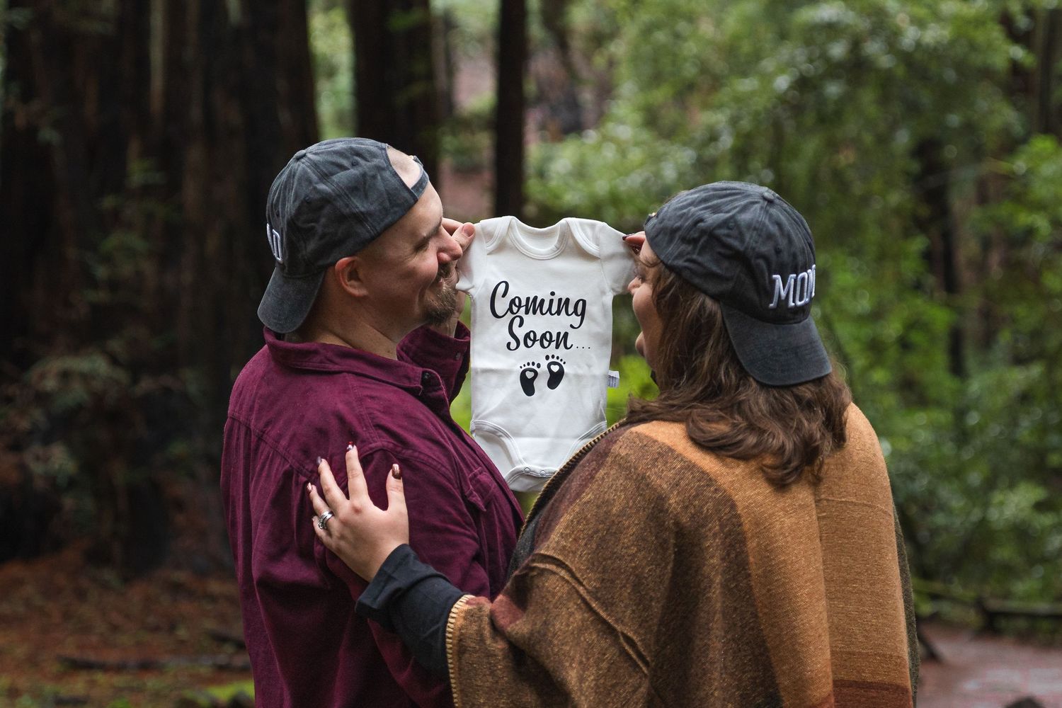 Couple holding a Coming Soon baby onesie in a forest, announcing their pregnancy.