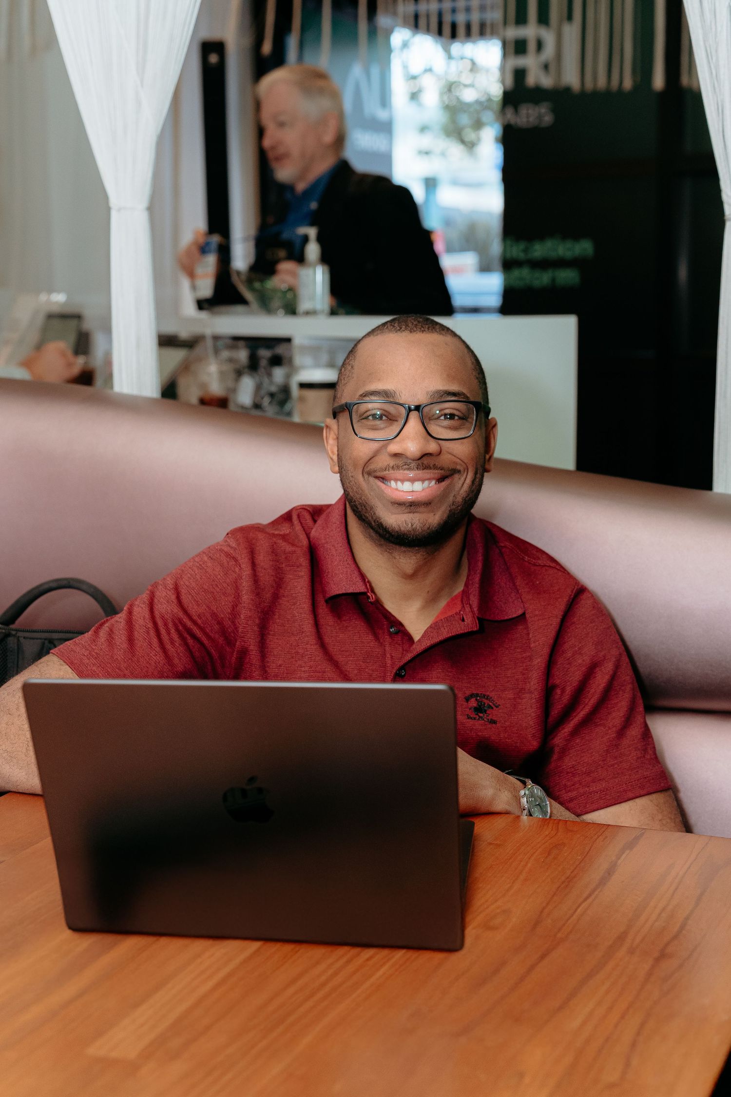 Smiling man in red polo shirt working on MacBook laptop at wooden table in modern cafe setting.
