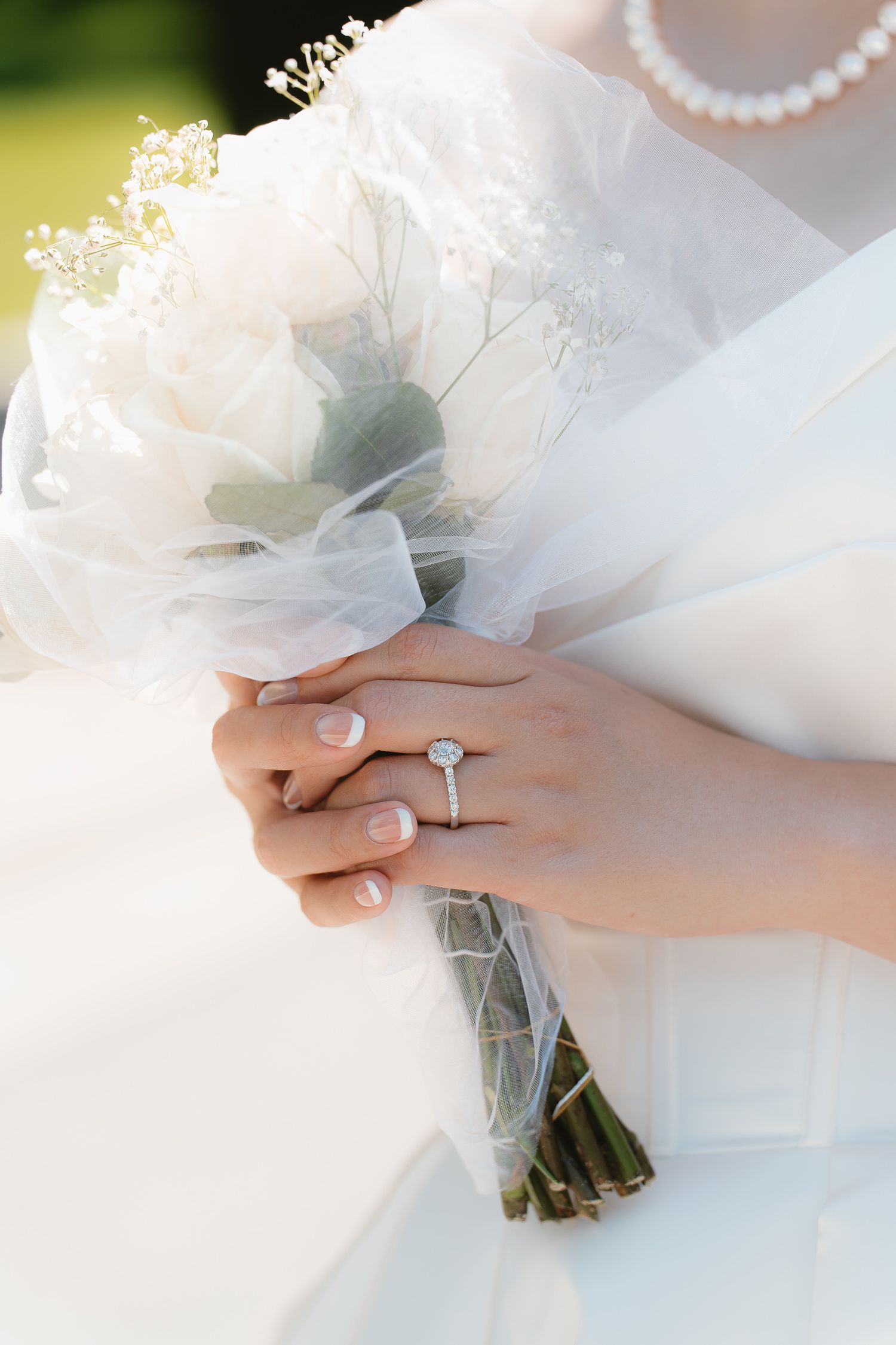 Bride holding white rose bouquet with baby's breath, showcasing diamond engagement ring and pearl necklace in elegant wedding attire.