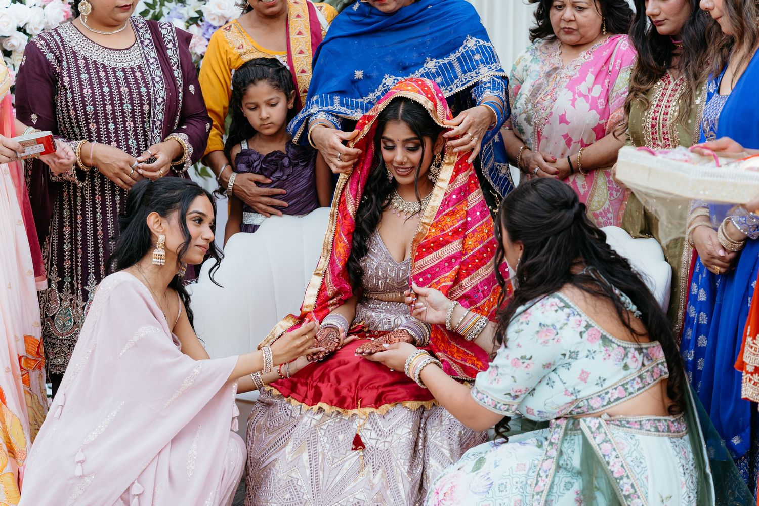 Indian bride in red lehenga surrounded by bridesmaids in pink during traditional wedding ceremony.