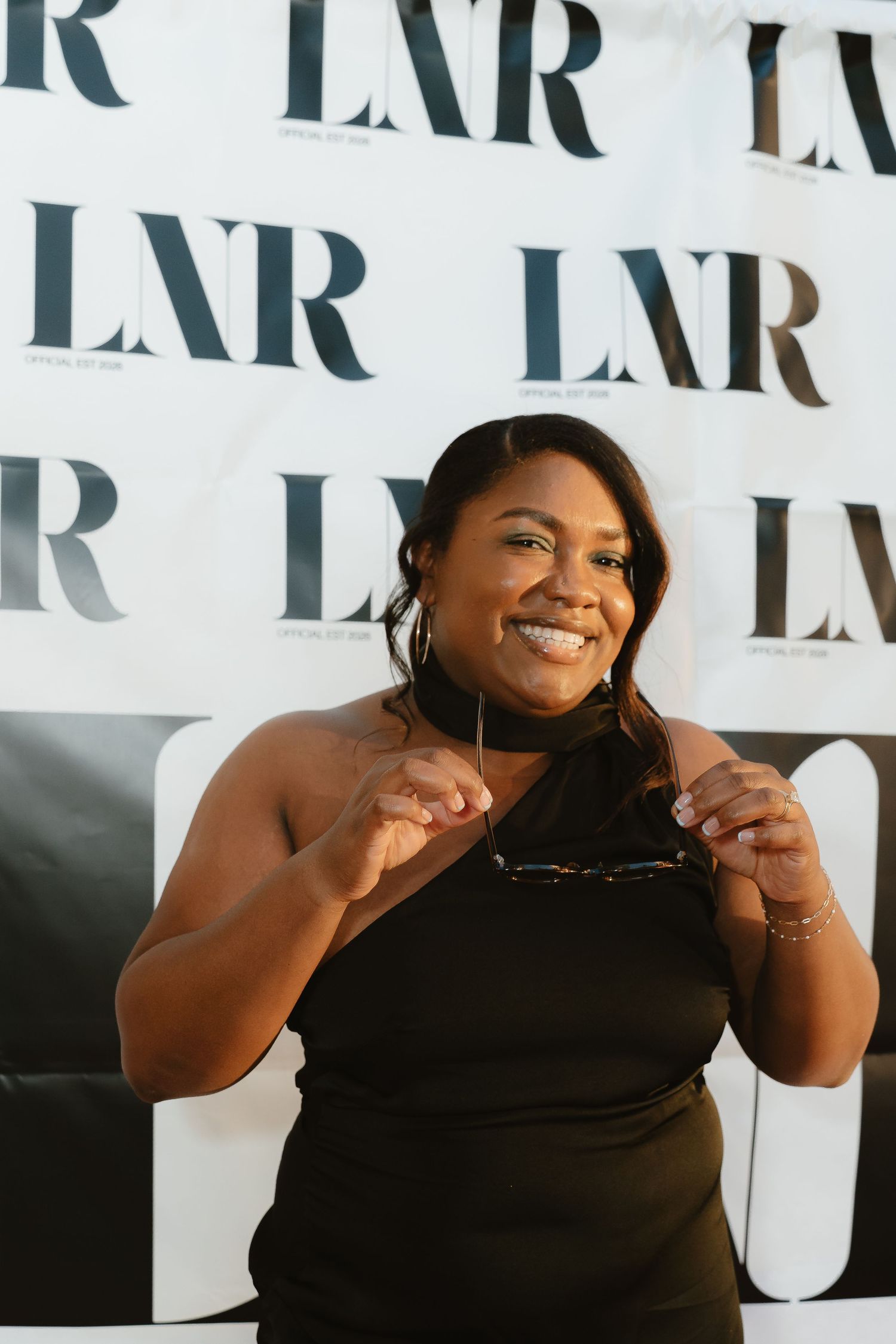 Smiling woman in black one-shoulder dress poses at LNR branded backdrop, holding sunglasses at an event.