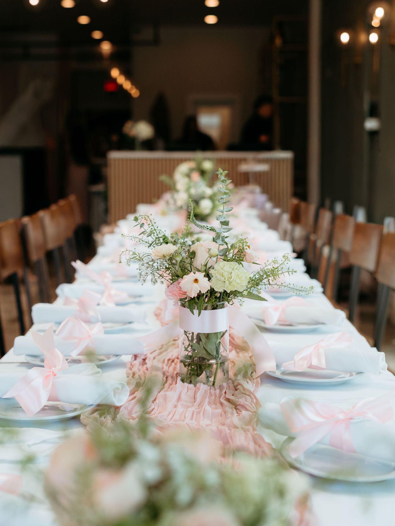 Elegant long reception table with pink bow napkins, floral centerpieces, and string lights in a modern venue.
