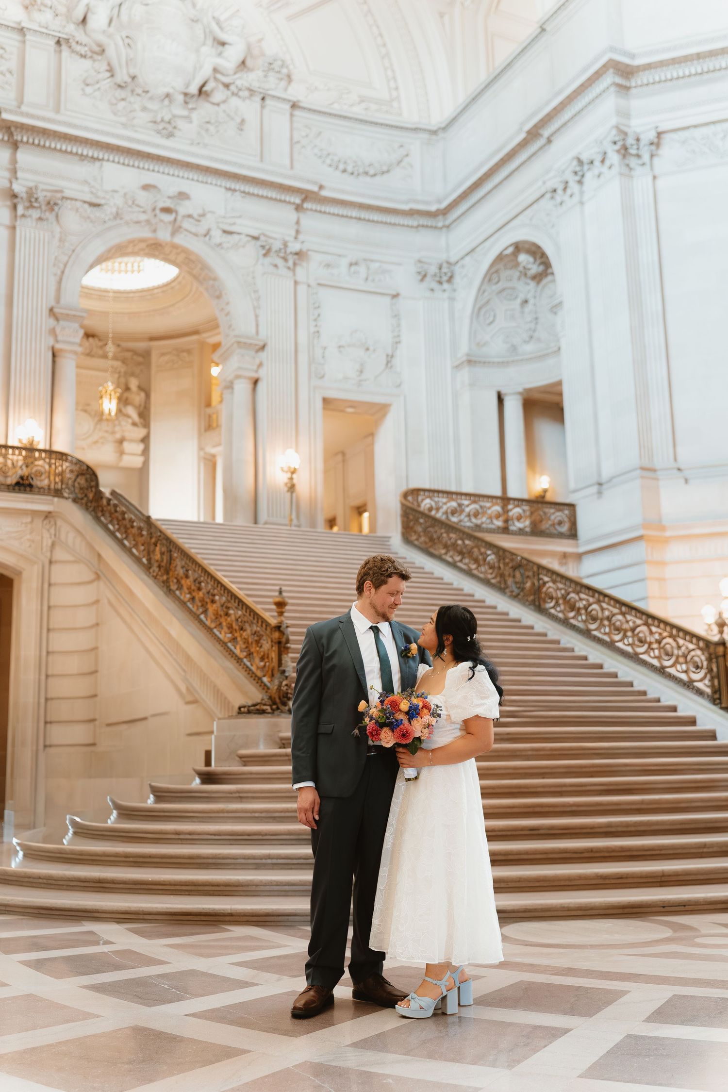 Couple poses at San Francisco City Hall grand staircase, bride in white dress with bouquet, groom in gray suit.
