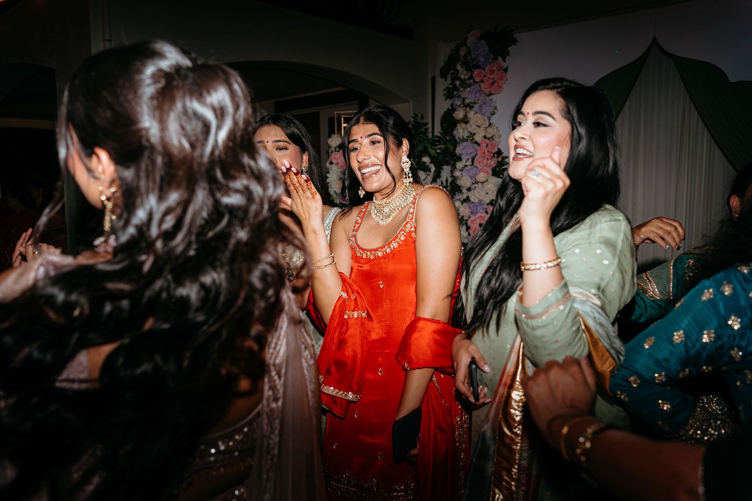Women in vibrant South Asian attire celebrate together at a festive wedding event, laughing and dancing joyfully.