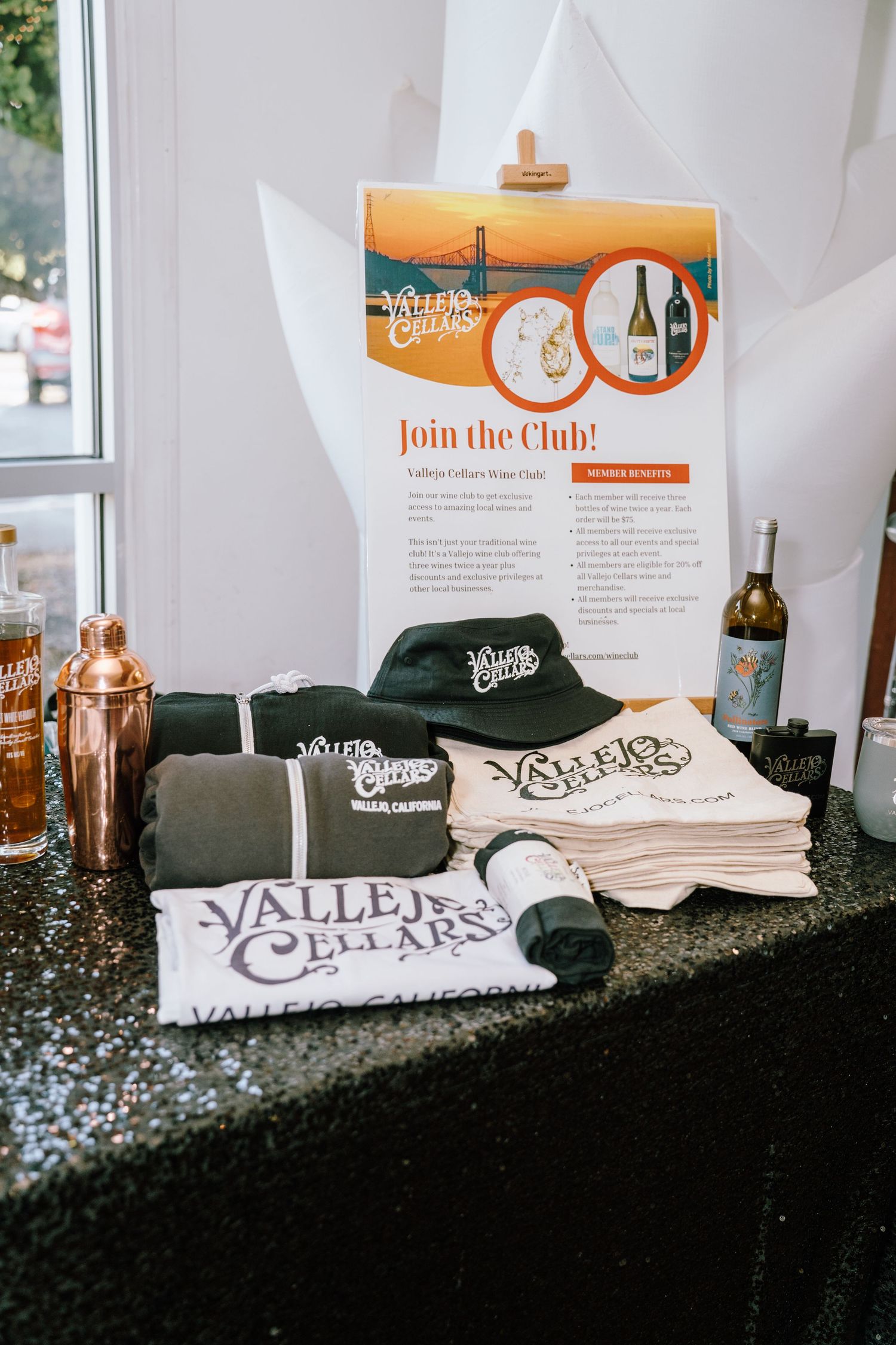 Vallejo Cellars wine club merchandise display featuring branded t-shirts, hats, and towels with a Join the Club promotional poster.