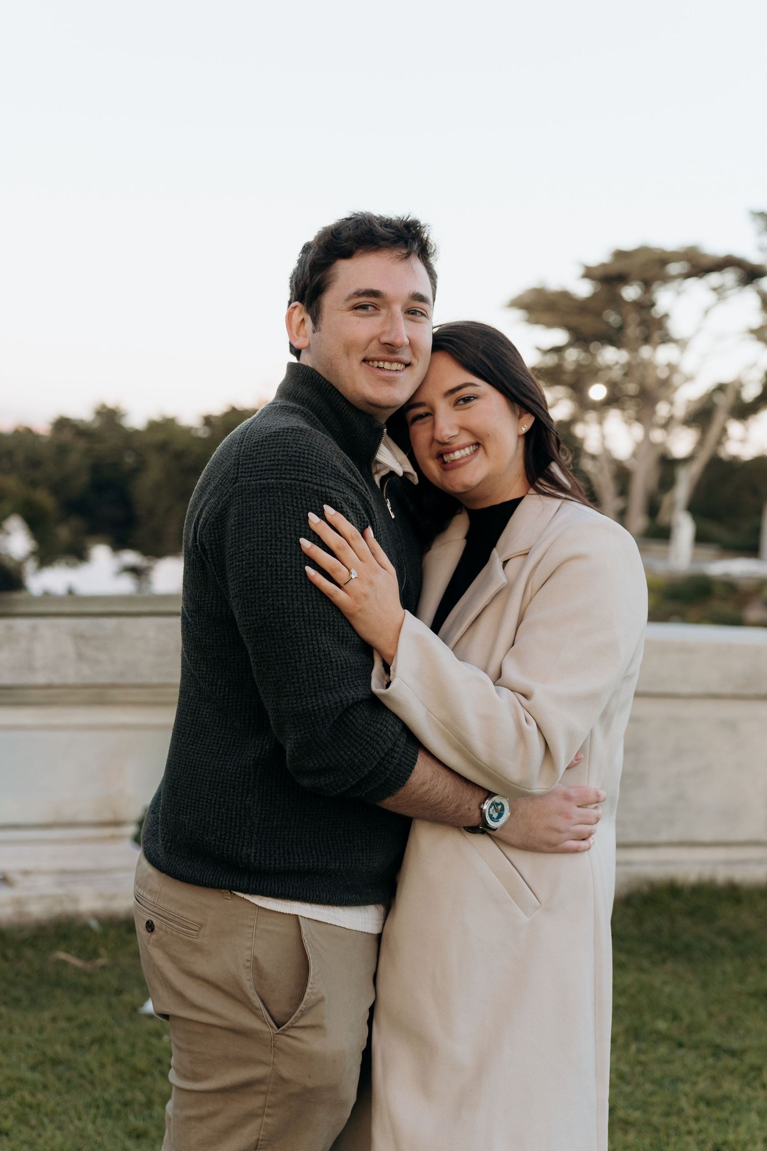 A smiling couple embraces outdoors, woman in beige coat and man in dark sweater, posing together in a park setting.