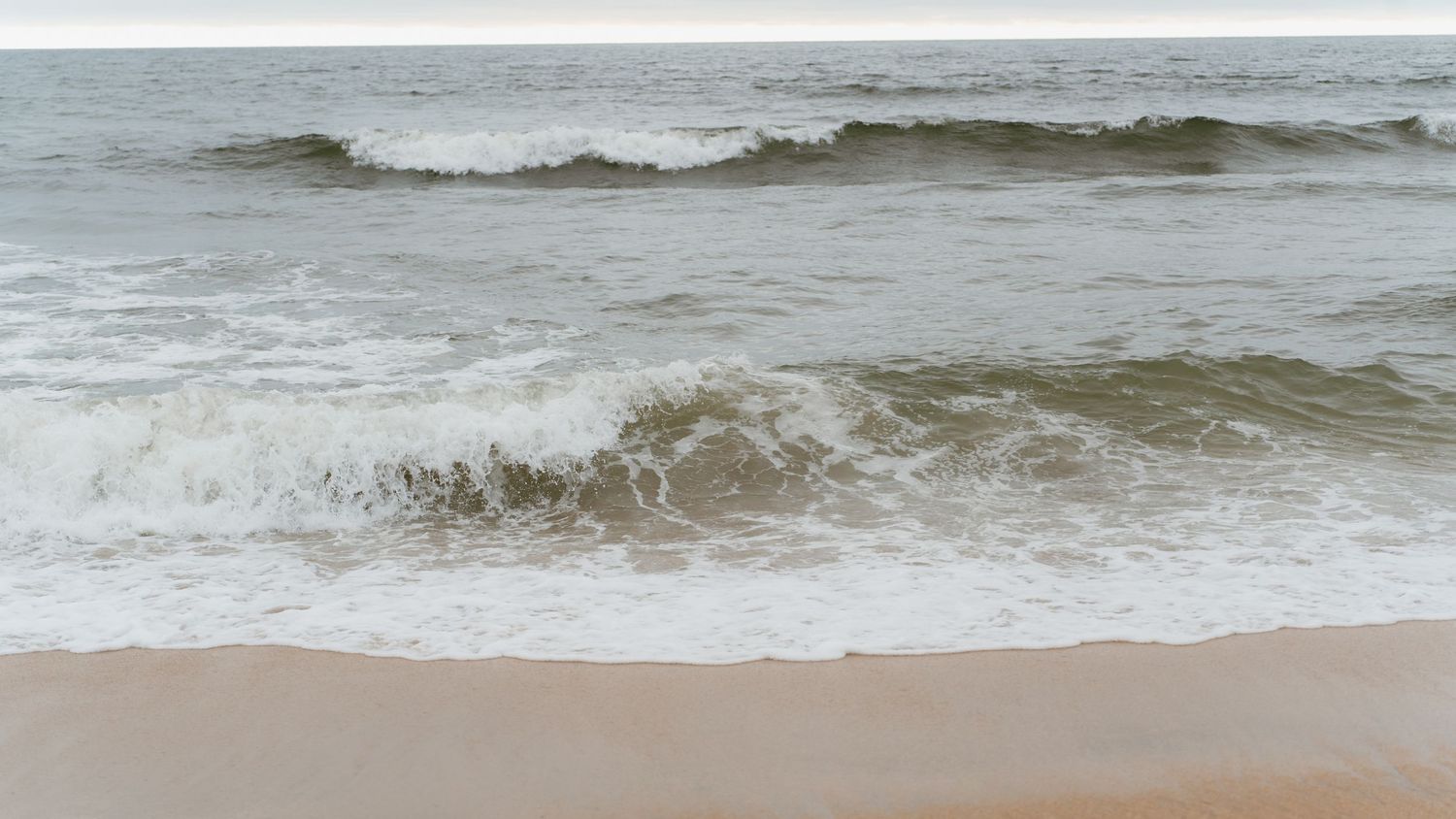 Gentle ocean waves washing onto a sandy beach shore with calm sea and overcast sky.