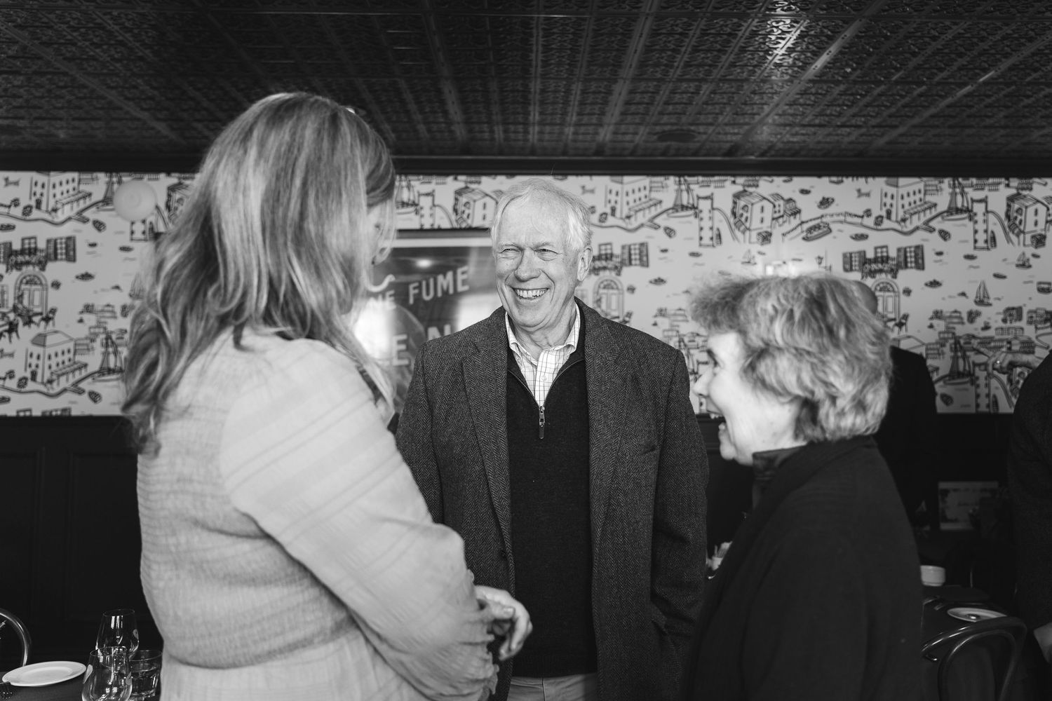 Black and white photo of three people smiling and conversing at an indoor social gathering.