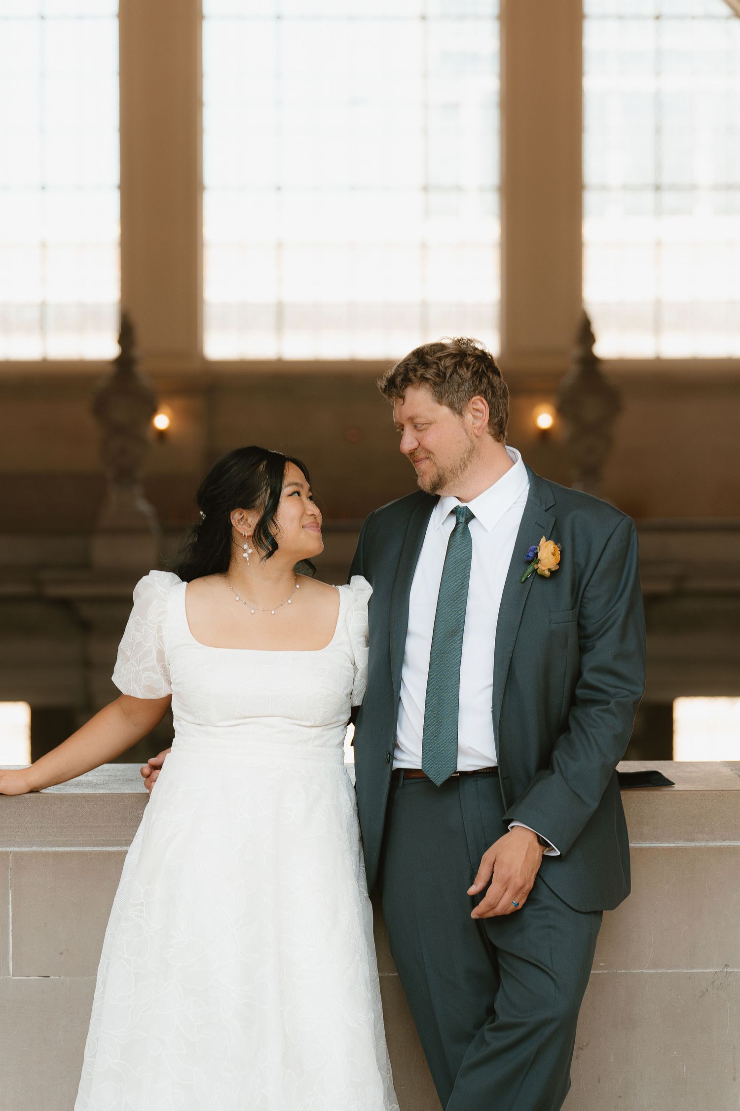 Bride and groom share a tender moment leaning against stone railing inside elegant wedding venue with tall windows.