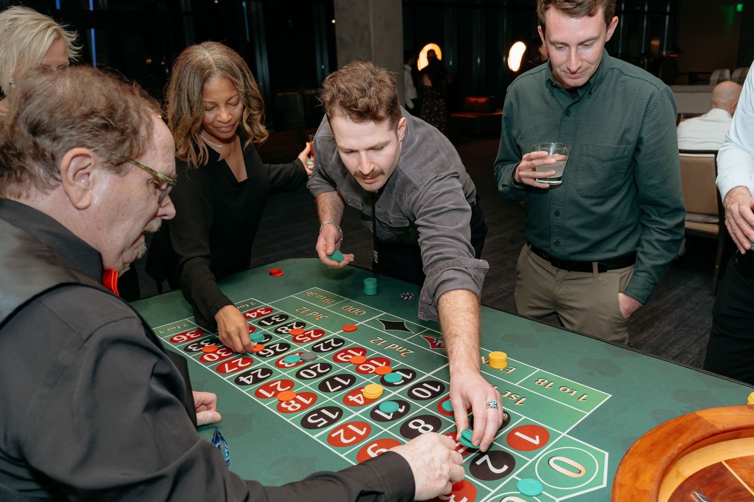 Group of people playing roulette at a casino table, placing bets on a green felt layout.
