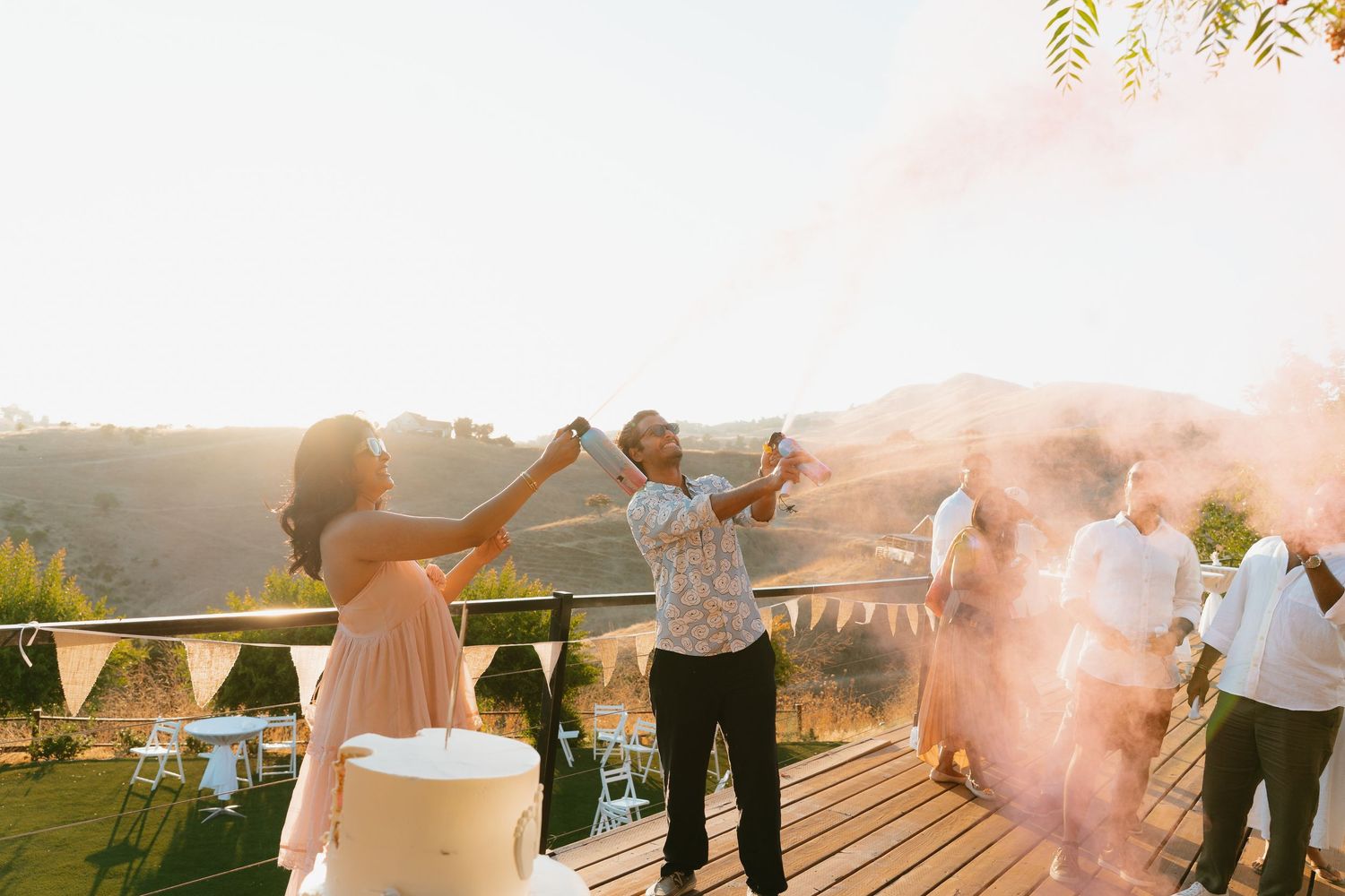 Guests celebrate outdoors with colorful smoke bombs at a rustic hillside wedding reception.