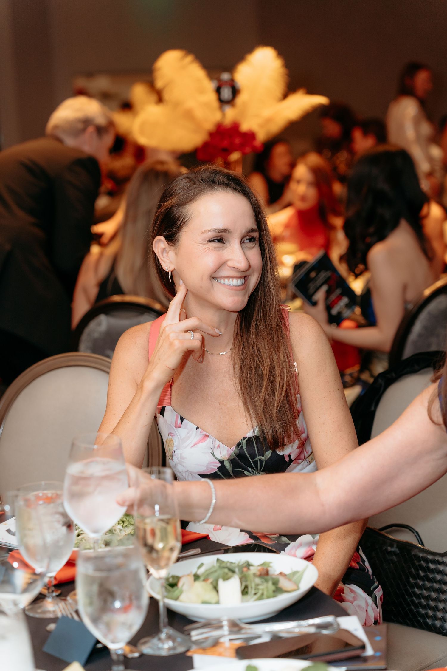 Smiling woman in floral dress seated at elegant gala dinner table with wine glasses and salad plate.