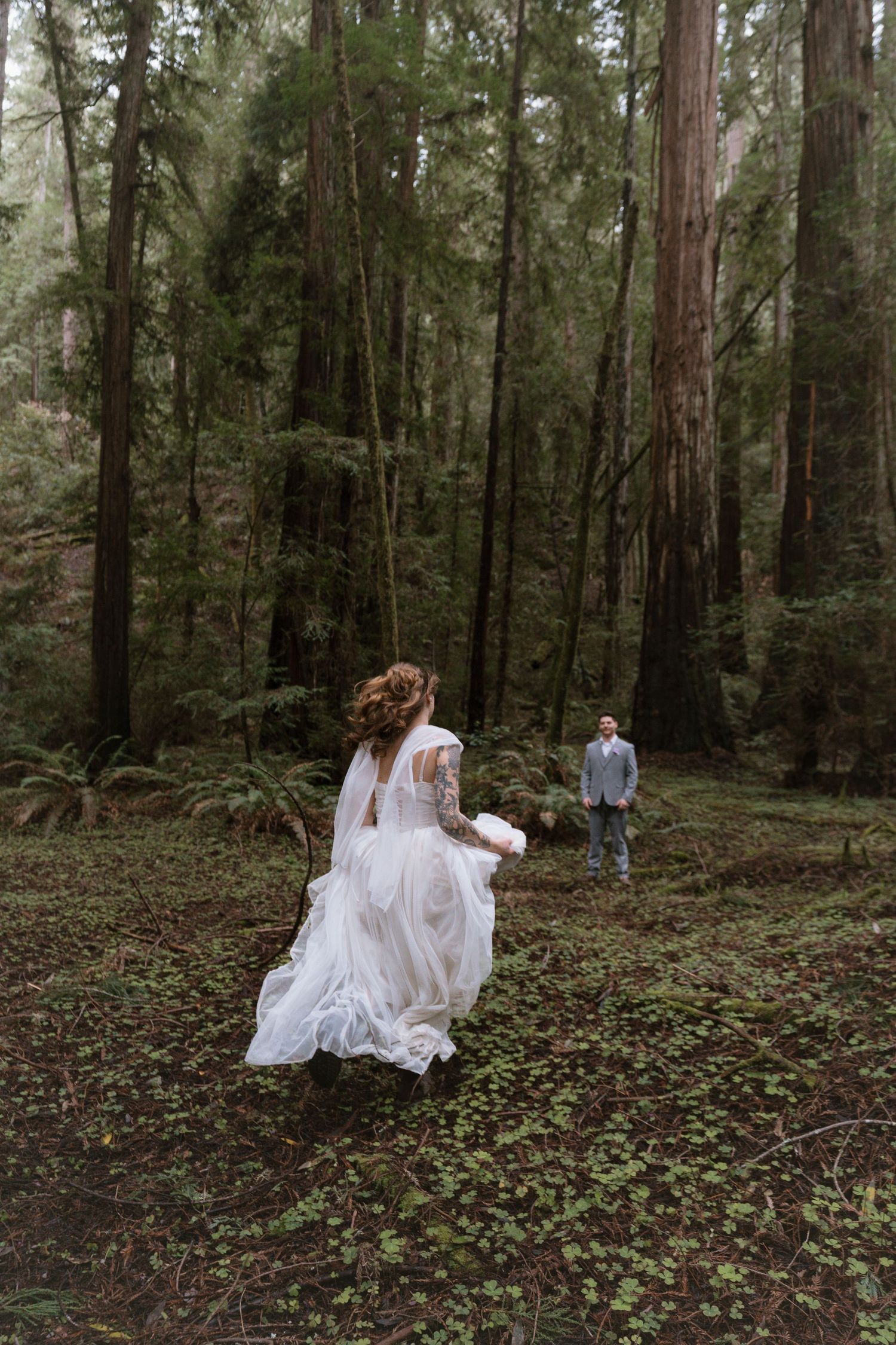 Bride in white gown runs toward groom in misty redwood forest during romantic outdoor wedding ceremony.