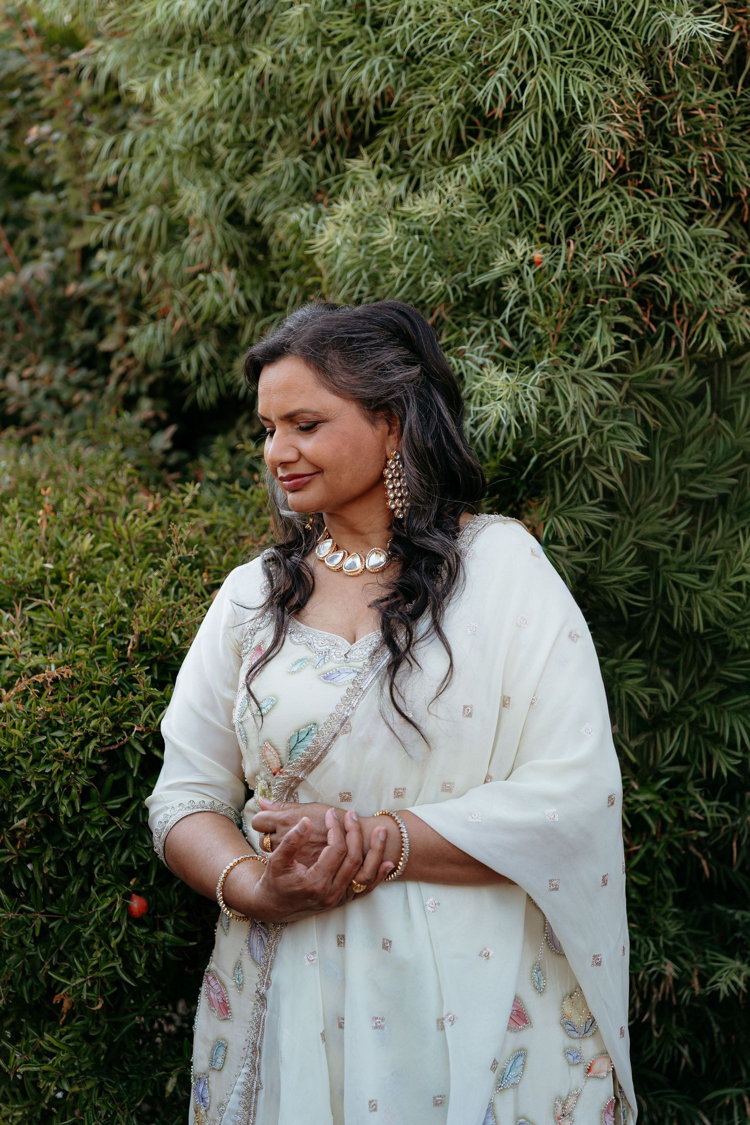 Woman in white embroidered salwar kameez with gold jewelry smiling outdoors near green foliage.