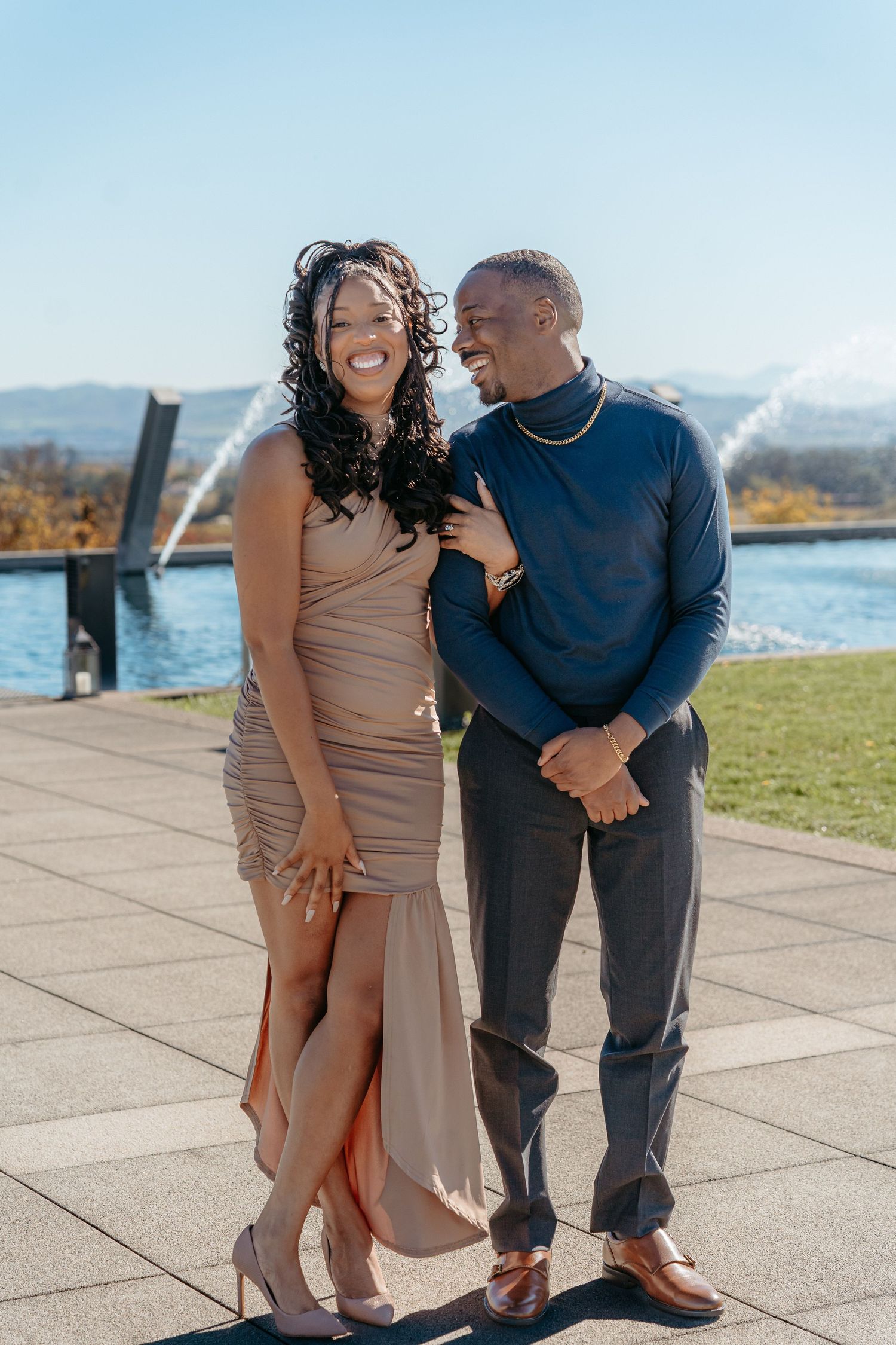 A joyful couple poses outdoors near a fountain, woman in beige dress and man in navy turtleneck, smiling together on a sunny day.