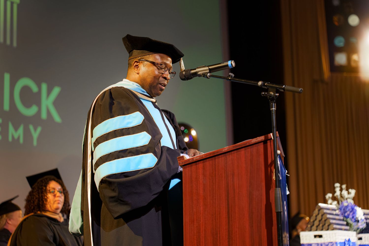Academic speaker in doctoral regalia delivers address at podium during formal graduation ceremony.
