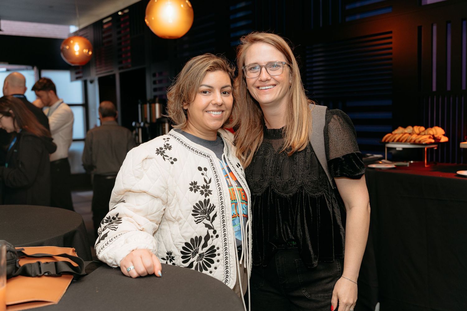 Two women smiling together at an indoor social event, one in a floral jacket and one in a black dress.
