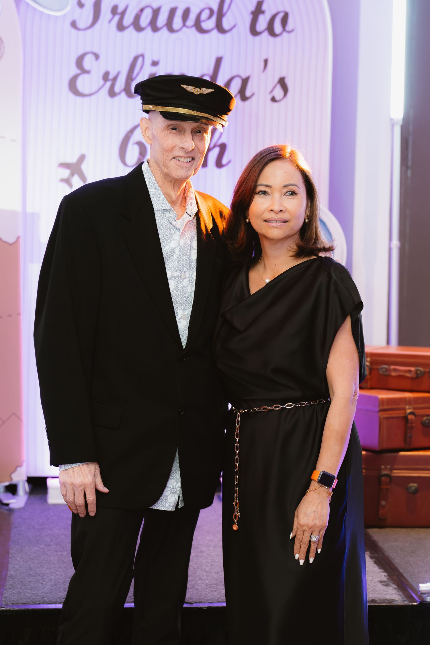 Couple posing together at Erlinda's 60th birthday travel-themed party, man wearing pilot hat, woman in black dress.
