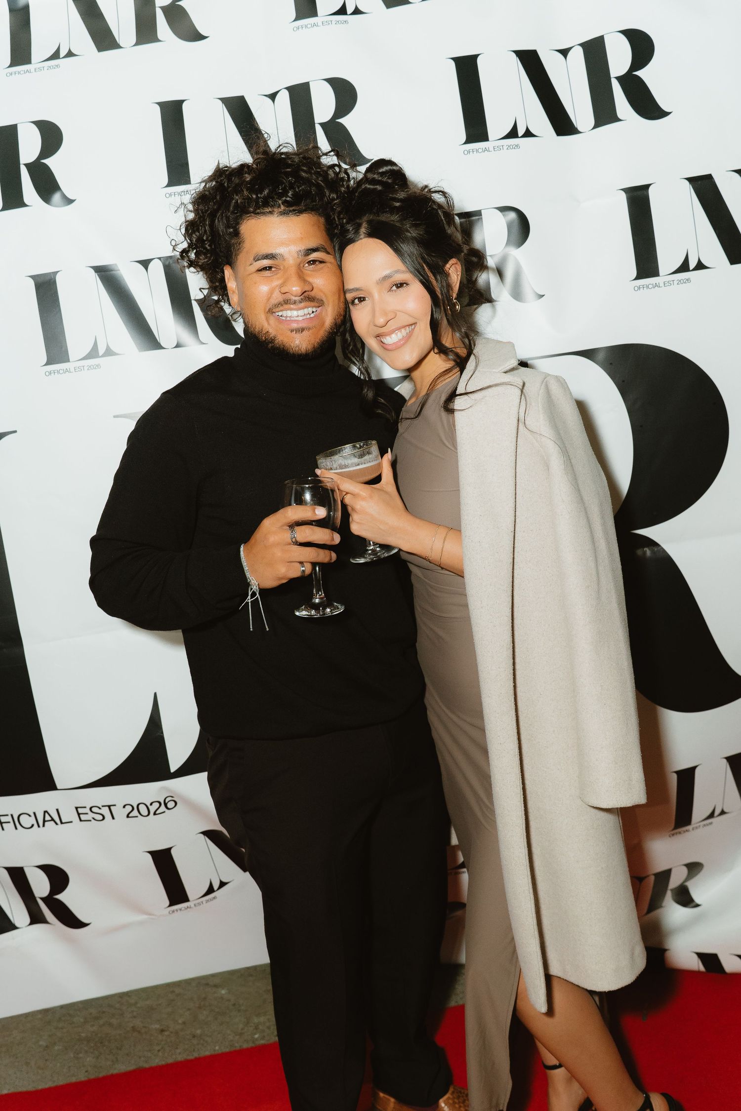 Stylish couple posing with drinks on red carpet at LXR magazine launch event.