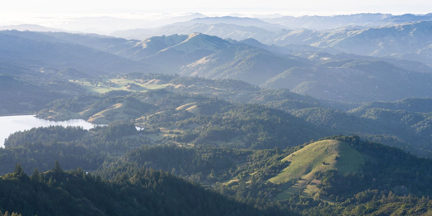 Aerial panoramic view of rolling forested hills and a small lake in the California coastal mountains.