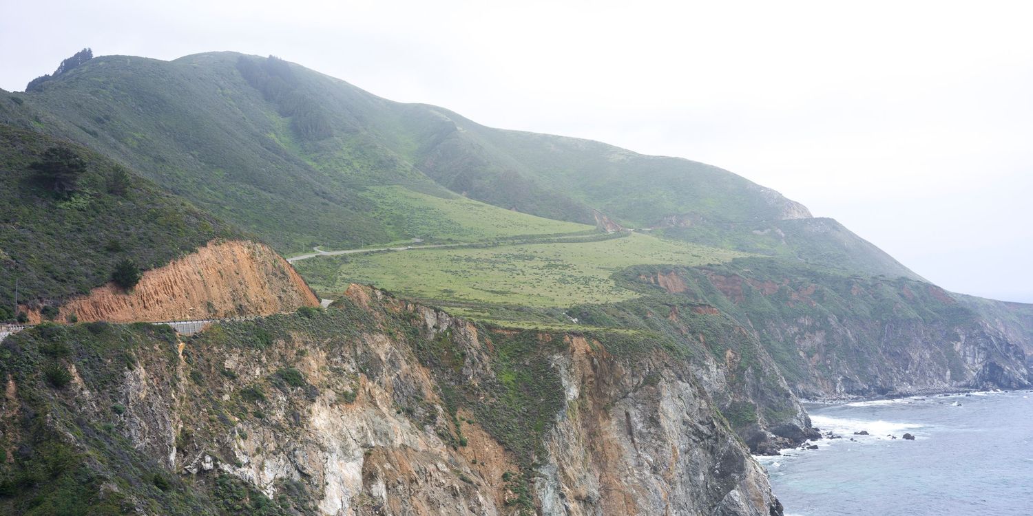 Steep green coastal cliffs and winding road along the dramatic Big Sur coastline in California.
