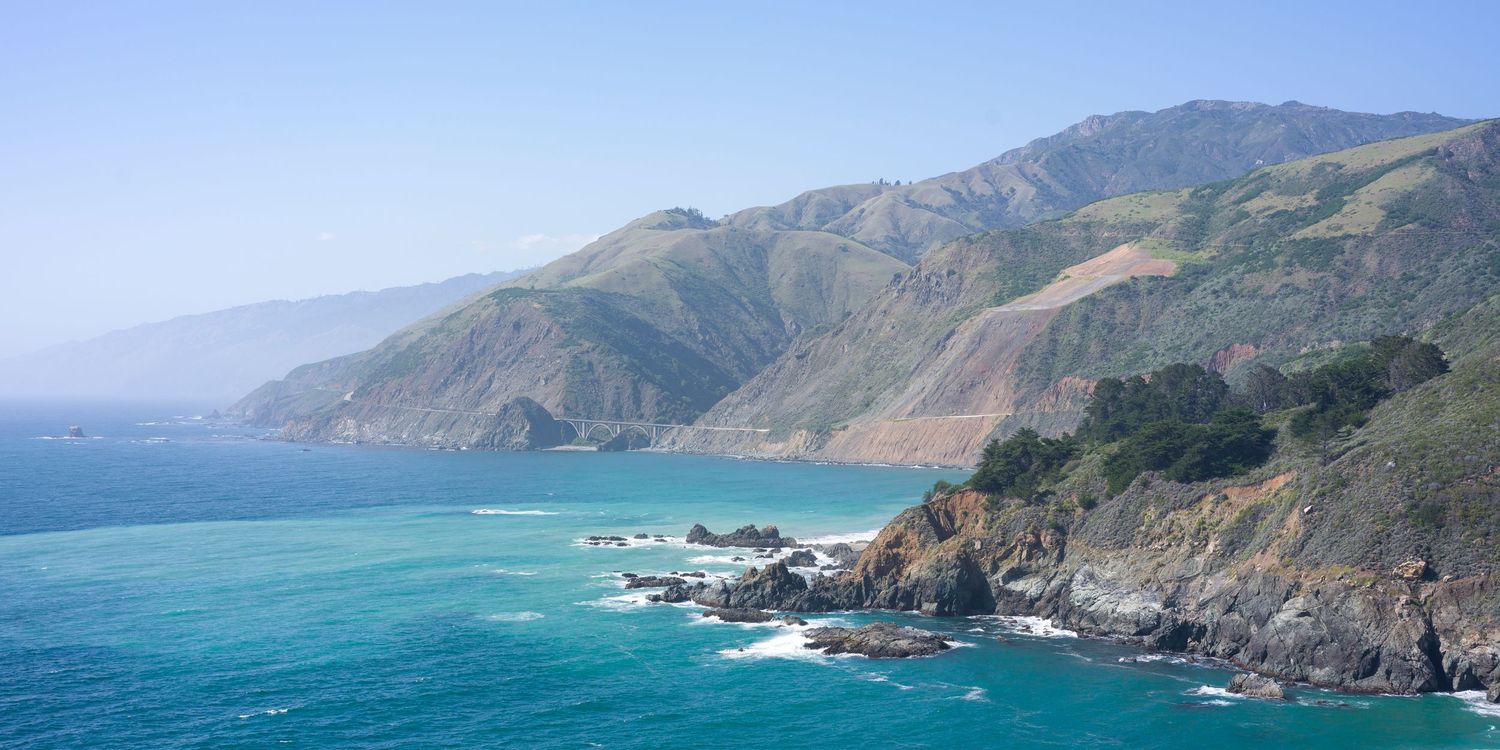 Turquoise waters of Big Sur coastline with rugged mountains and rocky shores on a clear day.