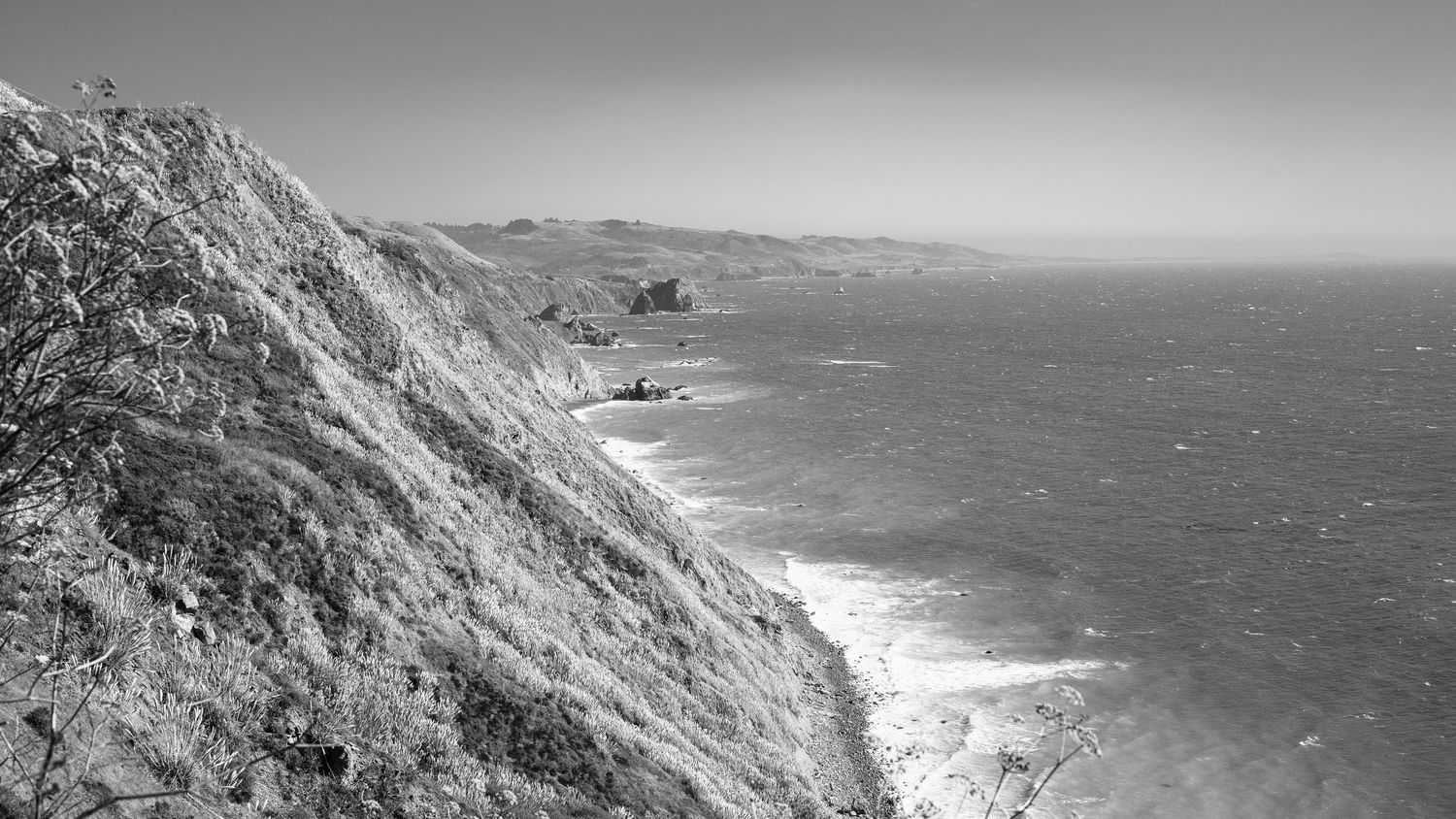Black and white coastal landscape featuring dramatic cliffs overlooking a vast ocean shoreline.